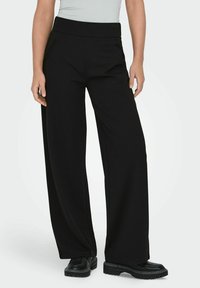 Osoba w czarnych, szerokich spodniach z bocznymi kieszeniami oraz czarnych, masywnych butach, stojąca na jednolitym tle.