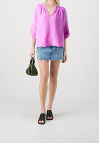 Blusa leggera rosa con scollatura a V e maniche a sbuffo, abbinata a una minigonna di jeans blu con orlo sfilacciato, sandali neri a scivolo e una borsa verde.