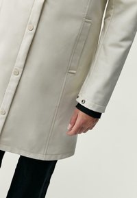 Béžový nepremokavý kabát s hladkou textúrou, ktorý má zapínanie na gombíky, bočné vrecká a cvoky na manžetách.