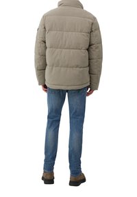 Mann in beiger Daunenjacke, blauen Jeans und braunen Schuhen, steht mit dem Rücken zur Kamera vor einem weißen Hintergrund.
