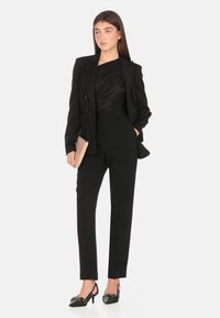 Completo nero su misura con giacca aderente, pantaloni plissettati e top nero arricciato. Abbinato a tacchi neri e una pochette color carne. Texture liscia.