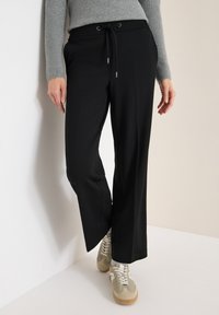 Zwarte wijde broek met een elastische tailleband en trekkoord, met mooie zijnaad en subtiele glans. G gedragen met beige sneakers.