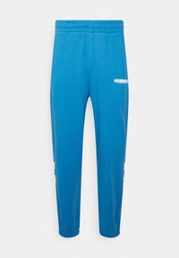 Blauwe sweatpants van zacht materiaal, met een elastische tailleband, taps toelopende pijpen en het witte "HUMMEL" logo op de dij.