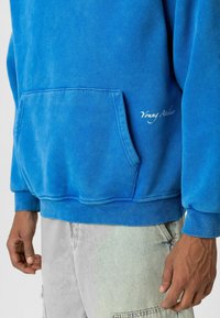 Blauwe sweatshirt met een buidelzak en geribbelde boord. Bevat het logo "Young Atelier" in het wit aan de onderkant rechts. Zachte stofstructuur.