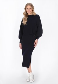 Granatowy sweter dzianinowy z balonowymi rękawami i wysokim dekoltem, zestawiony z dopasowaną spódnicą midi z bocznym rozcięciem oraz białymi sportowymi butami za kostkę.