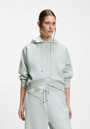 Frau trägt einen hell pastellblauen Hoodie mit Kordeln und passende Jogginghose, steht vor einem schlichten weißen Hintergrund.