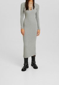Graues, geripptes Strickkleid mit langen Ärmeln, tiefem V-Ausschnitt und Ausschnittdetail. Kombiniert mit schwarzen Leder-Stiefeletten mit silberner Hardware.