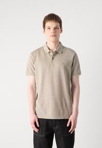 Beige polo skjorte med korte ermer, krage, og subtile stripestriper ved kantene. Har en liten logo på brystet. Brukt med mørke jeans.