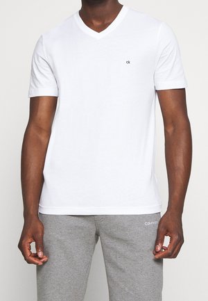 Homme portant un t-shirt blanc uni à col en V avec un petit logo "ck" sur la poitrine et un pantalon de survêtement gris avec le texte blanc "Calvin Klein" sur la cuisse.
