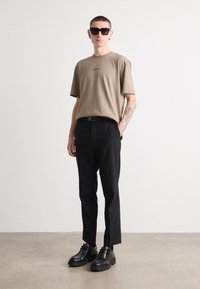Beige pamut póló logóval, fekete zakózott nadrág, fekete cipő és négyzet alakú napszemüveg. Simaság jellemzi, minimalista dizájn.