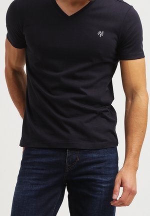 T-shirt noir en coton à col en V et manches courtes, avec un petit logo blanc sur la poitrine. Porté avec un jean bleu foncé.