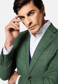 Zielona marynarka z tkaniny w typie tweed, w wzór w jodełkę, z wyrazistym kołnierzem i małym logo na lewym klapie, noszona na białej koszuli.