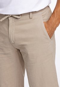 Beige linnebyxor med avslappnad passform, med dragsko i midjan, sidofickor och subtil textur. Sömmarna är synliga.