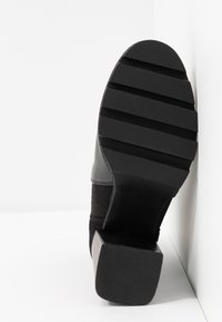 Musta korkonilkkuri, jossa on teksturoitu nahka- ja kangaspäällinen. Pohjassa on syvät urat pitoon ja suorakaiteen muotoinen korkopohja.