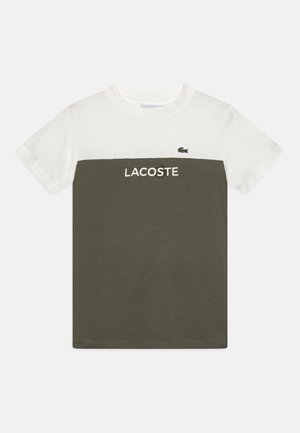 Koszulka z krótkim rękawem z białą górną częścią i oliwkowo zieloną dolną, z napisem „LACOSTE” i małym logo krokodyla na piersi.