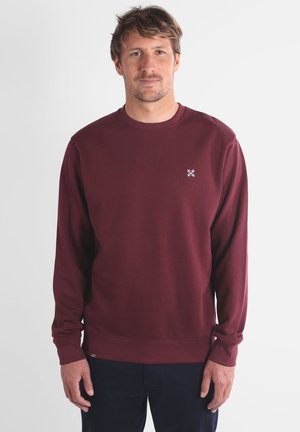 Sweatshirt col rond bordeaux en tissu doux, avec un petit logo blanc sur la poitrine et des poignets et un ourlet côtelés.