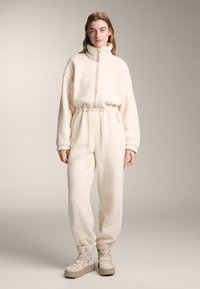 Bela, puhasta flis kombinezon z visokim ovratnikom, nastavljivim pasom in manšetami na gležnjih, kombiniran z bež nizkimi čevlji na platformo z zavezovanjem.