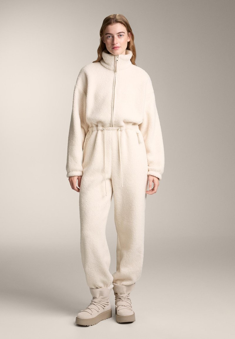 Weißer, flauschiger Fleece-Overall mit hohem Kragen, verstellbarer Taille und Bündchen an den Knöcheln, kombiniert mit beigen, geschnürten Plateaustiefeln.