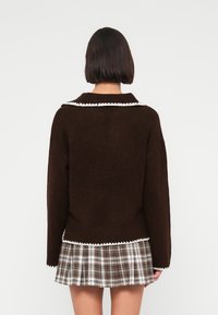 Καφέ πουλόβερ με λευκή σκαλοπατιαστή γιακά· απαλή υφή. Συνδυάζεται με πλισέ καρό φούστα σε καφέ και λευκούς τόνους.