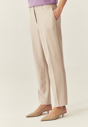 Osoba v béžových na mieru šitých nohaviciach s bočnými vreckami a béžových lodičkách na nízkom podpätku so špičatou špičkou, stojaca na jednoduchom pozadí.