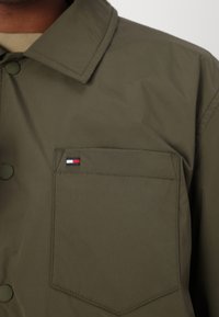 Olivengrøn jakke lavet af blødt stof med spids krave, trykknapper og en brystlomme med et lille logo i rød, hvid og blå.