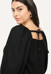 Žena s tmavými vlasy, která má na sobě zlaté kruhové náušnice a černý top s nafouknutými rukávy a zavázanou mašlí na zadním výstřihu.