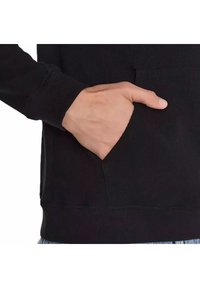 Sudadera negra con tejido texturizado, bolsillo canguro y puños ajustados, con una mano descansando en el bolsillo del lado derecho.