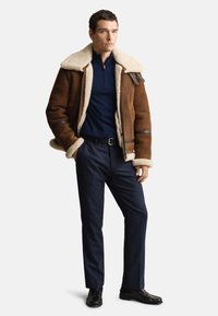 Bruine shearling jas met crèmekleurige voering, marineblauw overhemd en donkere broek. De jas heeft leren accenten en een ritssluiting.