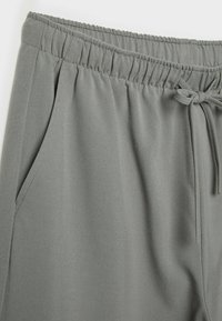 Pantalon gris avec taille élastique, poche latérale et cordon de serrage sur le devant côté droit.