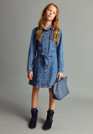 Vestito di jeans con maniche lunghe, chiusura a bottoni e vita fasciata; abbinato a stivali in camoscio blu navy e una piccola borsa blu. Texture morbida.