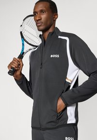 Casaco atlético preto com detalhes brancos e o logótipo "BOSS". Apresenta um fecho frontal com zip, punhos canelados e tecido texturizado. Modelo a segurar uma raquete de ténis.