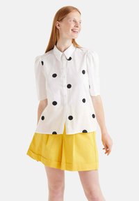 Camisa blanca de botones con lunares negros, mangas abullonadas y cuello clásico, combinada con una falda amarilla lisa y con pliegues.