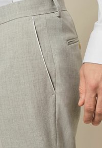 Pantalons gris clair en tissu texturé. Présentent une poche latérale avec un passepoil blanc et une petite poche à bouton à l'arrière.