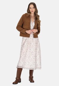 Giacca in suede marrone con due tasche frontali, indossata su un vestito bianco a fiori. L'outfit è completato da stivali marroni alti fino al ginocchio.