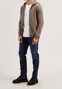 Man i brun zip-up hoodie, vit t-shirt, mörkblå jeans och svarta klumpiga skor, stående mot en enfärgad bakgrund.