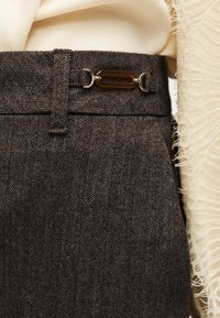 Gros plan d’un pantalon foncé à motif chevrons avec une boucle métallique ovale décorative sur la ceinture, associé à un tissu crème et à des détails en dentelle.