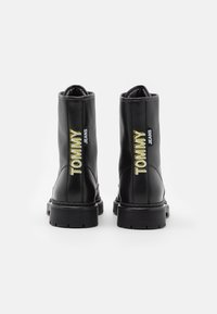 Botas de tobillo de cuero negro con textura estriada, suelas de goma anchas y bordado "TOMMY JEANS" en amarillo y blanco.