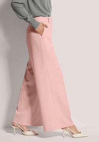 Wijde broek in zachtroze stof met een gladde textuur, voorzien van zijzakken en een op maat gemaakte tailleband, gecombineerd met witte hakken.