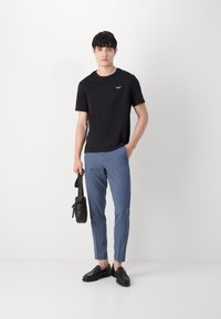 Zwarte korte mouw t-shirt met klein wit logo, lichtblauwe op maat gemaakte broek, zwarte instappers en een zwarte schoudertas.