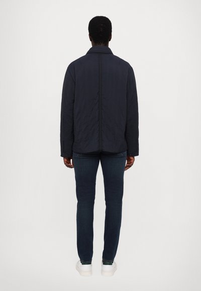 Navy-blau gesteppte Jacke mit Kragen, Knopfmanschetten und geradem Schnitt, kombiniert mit dunklen Jeans und weißen Sneakers.