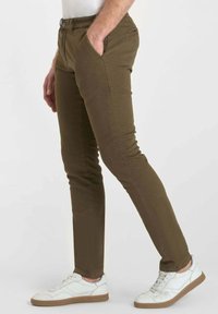Man in een slim-fit olijfgroene broek met zijn handen in zijn zakken en witte sneakers met een gumkleurige zool, staand tegen een effen achtergrond.