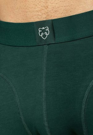 Lähikuva tummanvihreästä alusvaatevyötäröstä, jossa tikatut saumat ja pieni valkoinen lehtilogo keskellä edessä.