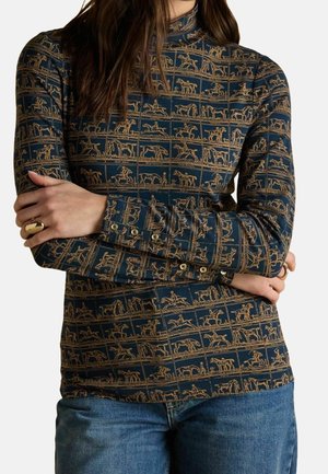 Kobieta ubrana w granatowy, długi rękaw z beżowym wzorem jeździeckim i niebieskie jeansy, z założonymi ramionami, ukazując złoty pierścionek na lewej dłoni.