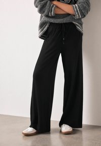 Black wide-leg pants feature an elastic waistband with a drawstring. Paired with white sneakers, showing a smooth fabric texture.