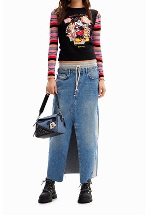 Kvinde iført langærmet top med striber og Mickey Mouse-tryk, blå denimnederdel med frontslids, sorte støvler og en håndtaske med Mickey Mouse-tema.