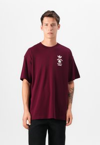 Burgundy cotton T-shirt with short sleeves, crew neck, and a white graphic design on the chest featuring brand logos and text.