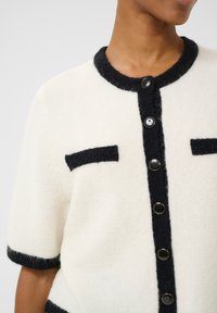 Short-sleeve cardigan in cream with black trim. Features black buttons and two black pockets. Soft texture, clean lines, and contrasting accents.