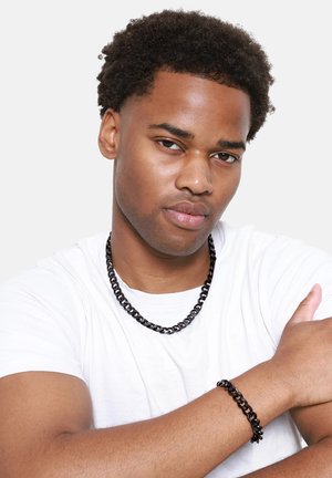 Schwarzes Kettenarmband und Halskette aus glattem Material, getragen von einer Person mit einem weißen T-Shirt vor einem neutralen Hintergrund.