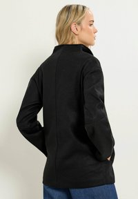 Svart, texturerad kappa med ståkrage och långa ärmar. Har en rak skärning och sidofickor, visas från bakvyn.