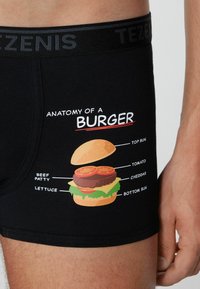 Boxer shorts negros con un gráfico colorido de la anatomía de una hamburguesa con ingredientes etiquetados: carne de res, lechuga, tomate, cheddar y panes.